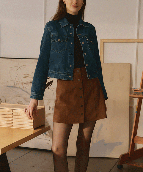 A man and a woman modeling casual apparel in a bright, modern art studio. The woman wears a blue denim jacket over a black top and brown skirt, while the man wears a brown button-up shirt and dark gray pants.