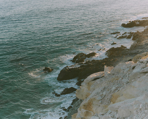 A scenic coastal view with turquoise ocean water, dark rocky shoreline, and pale beige cliffs under warm light.