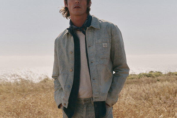 Man wearing a light denim jacket and scarf standing in a field with a blurred background