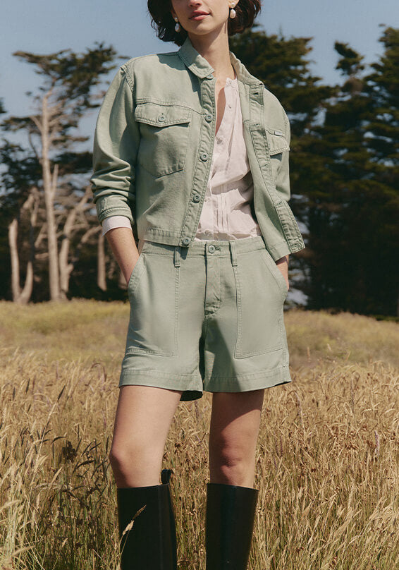 Person wearing a green jacket and shorts standing in a field with trees in the background