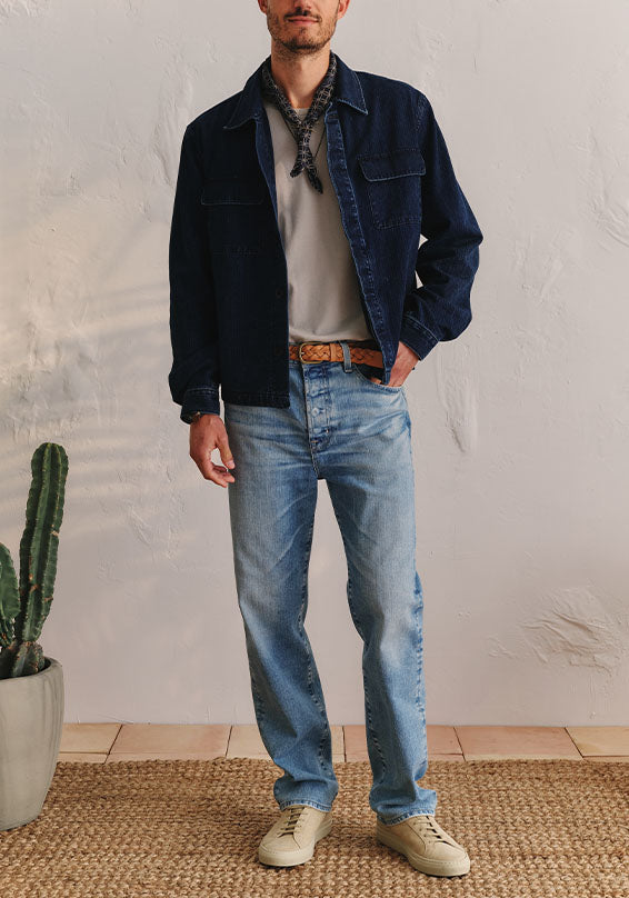 Man wearing a dark jacket, light shirt, blue jeans, and beige shoes standing against a white wall with a cactus plant on the left.