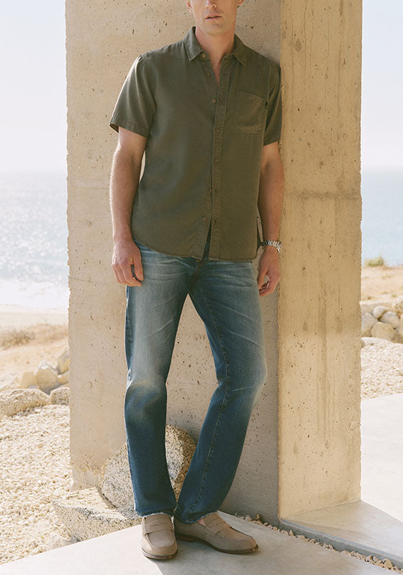 Man wearing a green shirt and blue jeans leaning against a stone wall.