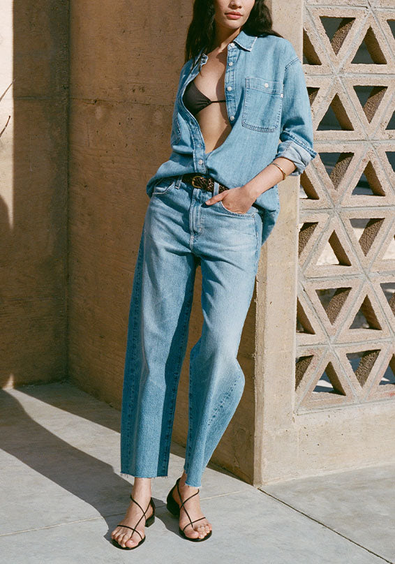 Woman wearing a denim shirt and jeans standing against a textured wall.