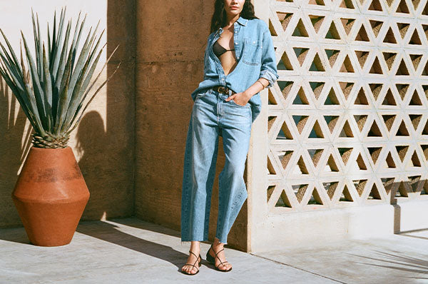 Woman in a denim outfit standing next to a geometric-patterned wall and potted plant.