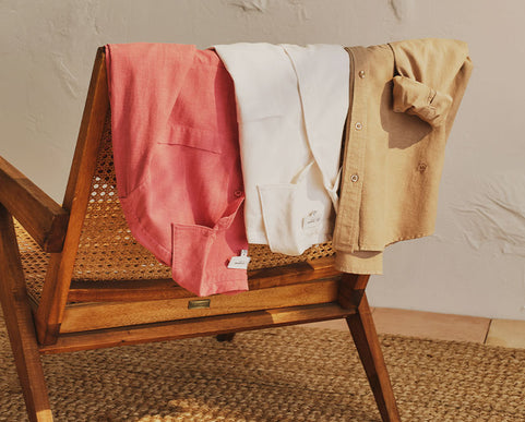 Three shirts in different colors draped over a wooden chair against a neutral wall.