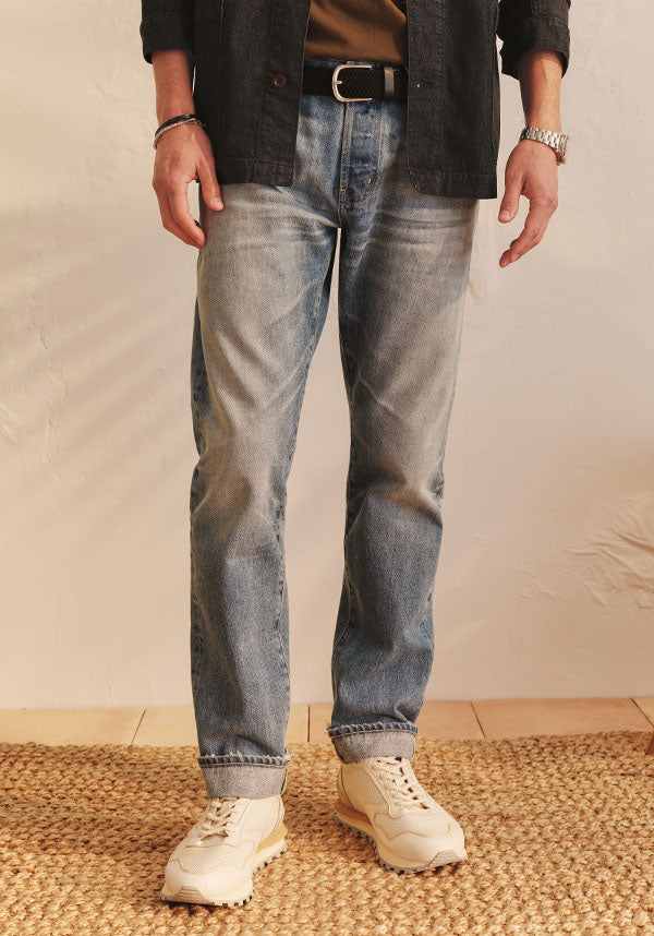 A person from the waist down wearing light-wash blue jeans with a black belt, a dark overshirt, and off-white sneakers, standing on a woven jute rug against a neutral beige wall.