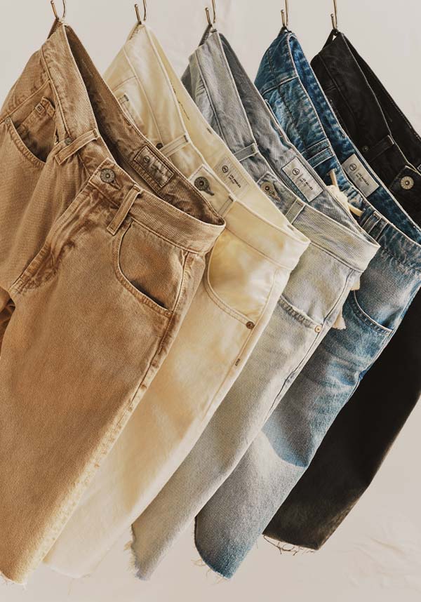 Five pairs of jeans in different washes hanging side by side against a plain light background. The colors include tan, cream, light gray, medium blue, and black.