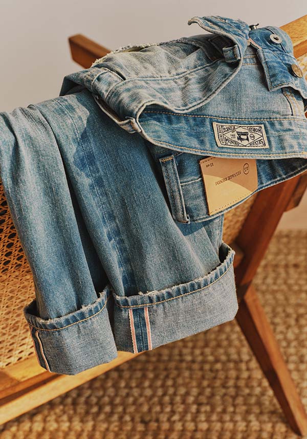 A pair of light blue denim jeans with rolled cuffs, draped over a wooden chair with a woven seat. A brand patch is visible on the back pocket, and a paper tag is tucked into the pocket.