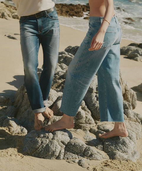 Two models wearing light blue denim jeans standing barefoot on rocks at a beach.