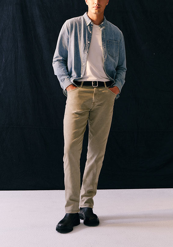 A man modeling a casual outfit consisting of a light blue denim button-up shirt, a white t-shirt, beige chinos, and black leather boots, standing against a black studio backdrop.