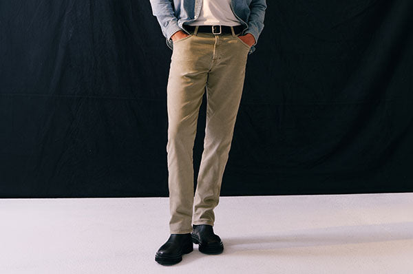 A man modeling a casual outfit consisting of a light blue denim button-up shirt, a white t-shirt, beige chinos, and black leather boots, standing against a black studio backdrop.
