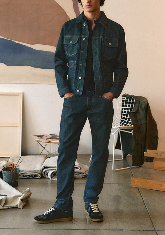A man wearing a dark blue denim jacket and matching dark blue jeans, paired with dark blue sneakers, standing in a minimalist studio setting with a painting and chair in the background.
