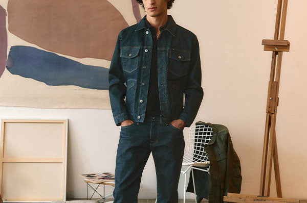 A man wearing a dark blue denim jacket and matching dark blue jeans, paired with dark blue sneakers, standing in a minimalist studio setting with a painting and chair in the background.