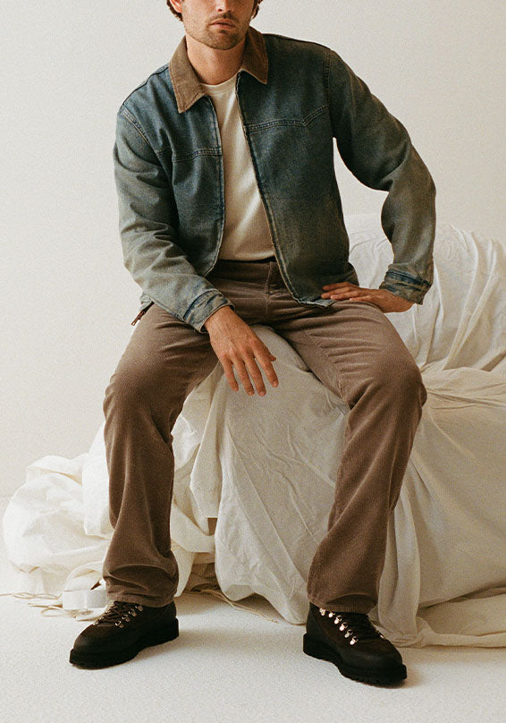 A man wearing a dark washed denim jacket with a brown corduroy collar, layered over a cream t-shirt and brown trousers, sitting on a white draped bed against a neutral studio background.