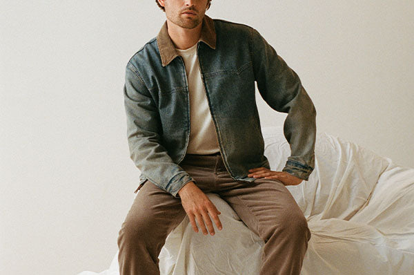 A man wearing a dark washed denim jacket with a brown corduroy collar, layered over a cream t-shirt and brown trousers, sitting on a white draped bed against a neutral studio background.