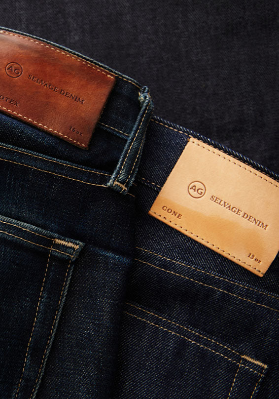 Close-up of dark indigo denim jeans showing two leather brand patches on the back waistband, one brown and one tan, both labeled AG Selvage Denim.