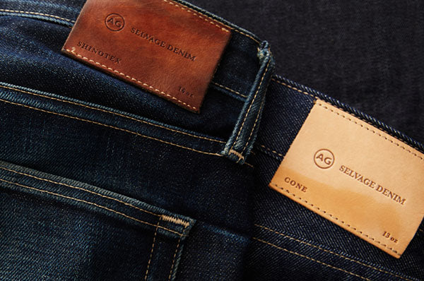 Close-up of dark indigo denim jeans showing two leather brand patches on the back waistband, one brown and one tan, both labeled AG Selvage Denim.