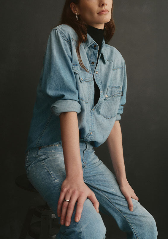 A woman wearing a light blue denim button-up shirt over a black turtleneck, paired with matching light blue denim jeans, posed against a dark gray studio background.