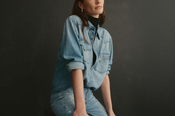 A woman wearing a light blue denim button-up shirt over a black turtleneck, paired with matching light blue denim jeans, posed against a dark gray studio background.