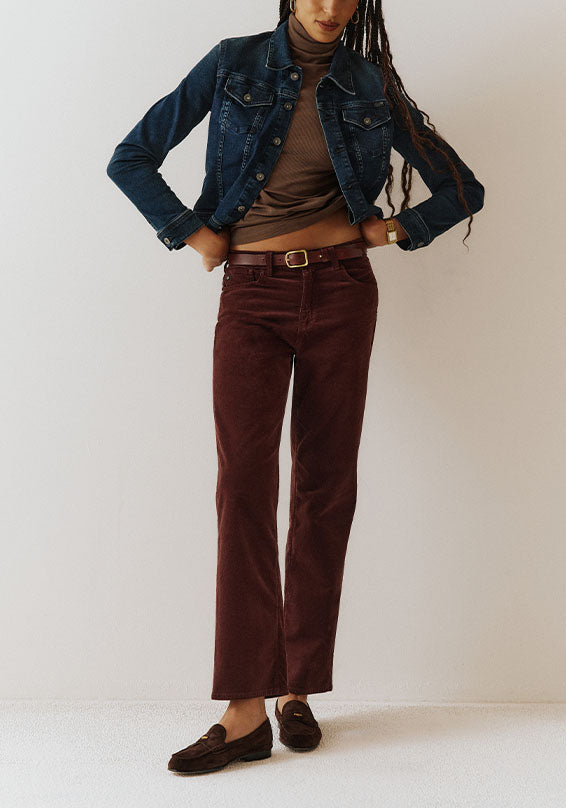 A model wearing a dark blue denim jacket, brown turtleneck, burgundy corduroy pants, and dark brown loafers, standing against a plain off-white studio background.