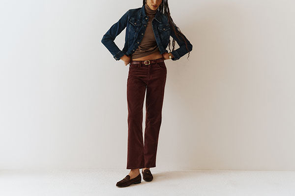 A model wearing a dark blue denim jacket, brown turtleneck, burgundy corduroy pants, and dark brown loafers, standing against a plain off-white studio background.