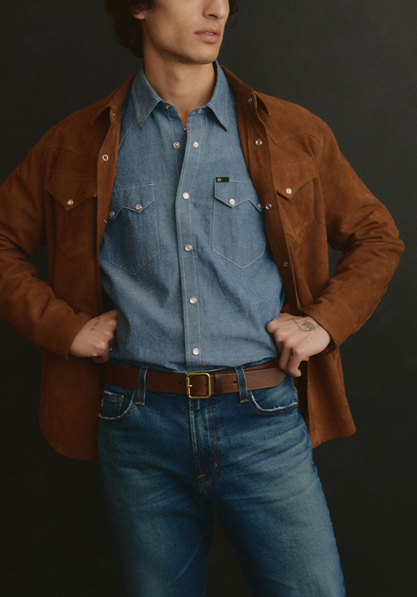 A man wearing a brown suede overshirt over a blue denim shirt and blue jeans, standing against a dark studio background.