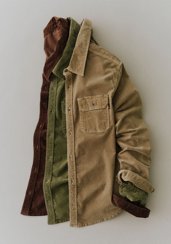 Three corduroy button-up shirts in brown, olive green, and tan are layered and displayed side-by-side against a neutral light gray background.