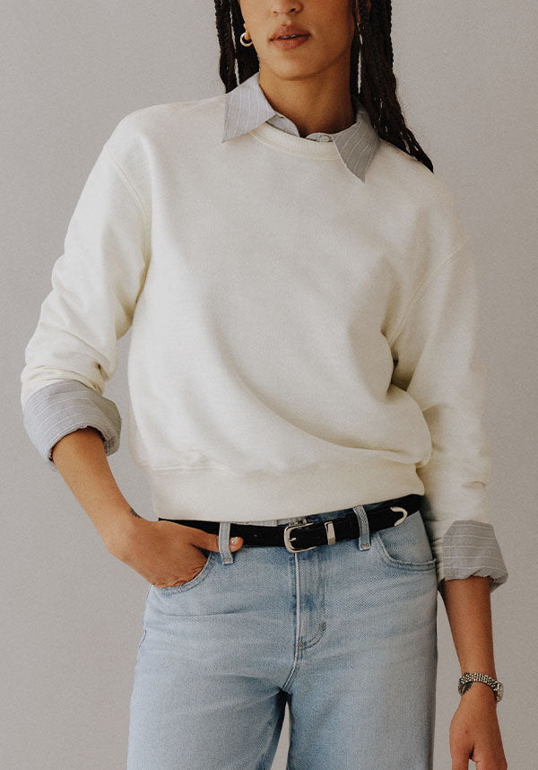 A person wearing a cream crewneck sweatshirt layered over a light gray striped button-down shirt, paired with light-wash blue jeans and a black belt, against a neutral gray background.