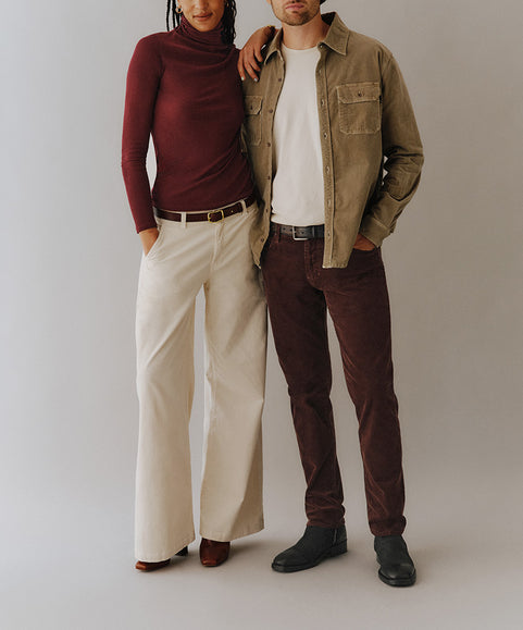 A man and a woman modeling coordinated outfits against a plain light gray studio background. The woman wears a burgundy turtleneck, cream wide-leg pants, and brown shoes.