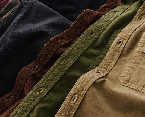 Close-up of folded button-front corduroy shirts in dark navy, brown, olive green, and tan, showing the textured corduroy fabric and snap-button placket.