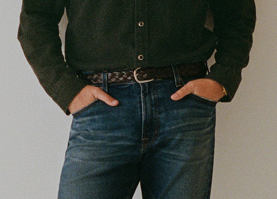 A person wearing dark blue jeans, a dark green button-up shirt, and a dark brown braided belt, standing with hands in pockets against a plain light gray background.