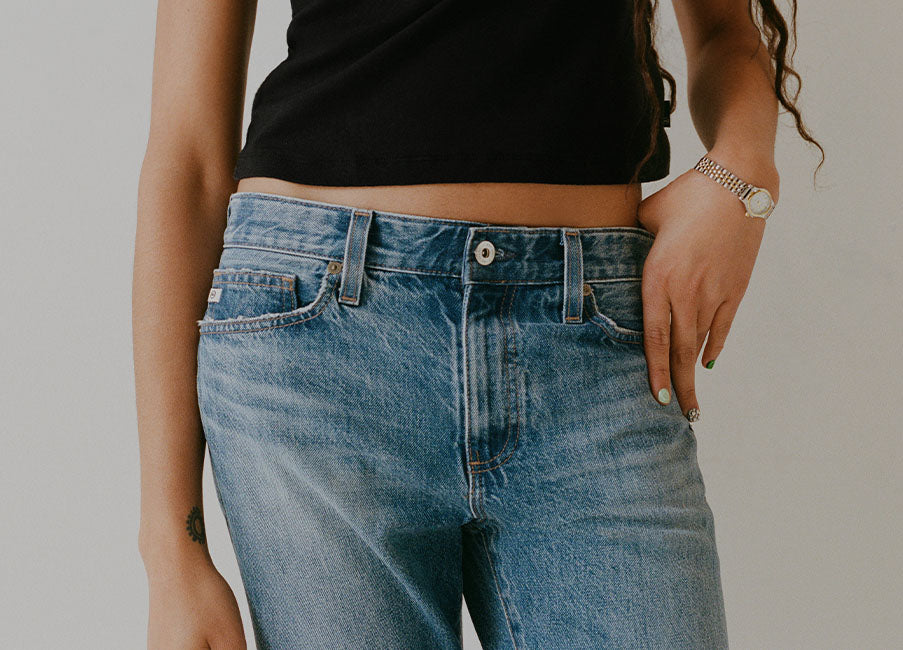 A close-up of a person wearing light blue denim jeans and a black cropped top, standing against a plain light gray background.